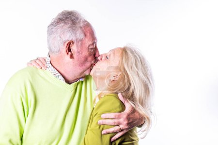 This heartwarming image captures a tender moment between a senior couple as they share a loving kiss. Both are dressed in vibrant sweaters, with a white background that accentuates the intimacy and