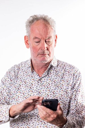 Ein Bild, das einen älteren Mann zeigt, der in die Nutzung seines Smartphones vertieft ist, zeigt einen modernen älteren Mann, der sich mit Technologie auskennt. Das florale Hemd verleiht dem krassen weißen Hintergrund einen Hauch von Lebendigkeit