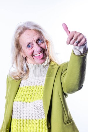 Eine überschwängliche Seniorin mit leuchtend blauen Augen streckt verspielt den Daumen in die Kamera, die Zunge in einer fröhlichen Geste herausgestreckt. Bekleidet mit einem lindgrünen Blazer über einem kuscheligen cremefarbenen Pullover und einem gelben