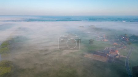 This image offers an aerial view of a misty countryside at sunrise. The soft morning light gently permeates through the veil of fog, revealing the outlines of the rural landscape below. The scattered