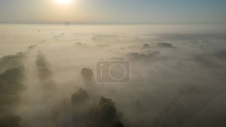 Diese Luftaufnahme zeigt eine atemberaubende Szene, in der ein frühmorgendlicher Nebel bei Sonnenaufgang einen dichten Wald umhüllt. Das Licht der aufgehenden Sonne filtert durch den Nebel und wirft einen warmen, goldenen Farbton