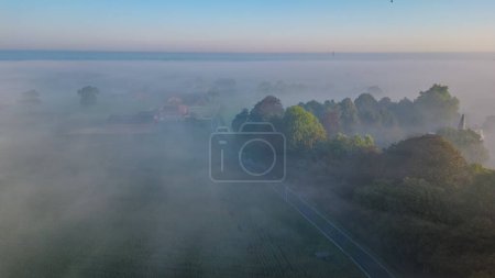 In this aerial photograph, a rural homestead emerges from the morning mist, with the first light of dawn casting a gentle glow on the scene. The homestead, nestled among sporadic trees and open