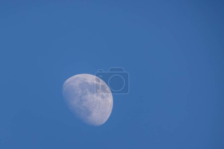 Esta imagen captura la fase gibbous encerada de la luna, claramente visible contra un cielo azul tranquilo, claro durante el día. La visibilidad de la superficie de las lunas detalles como cráteres, tierras altas