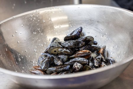 This image depicts a bowl of fresh, uncooked mussels still wet from washing. The stainless steel bowl reflects the light and features water droplets, emphasizing the cleanliness and freshness of the