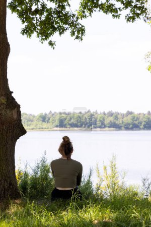 A serene image featuring a woman sitting beneath a leafy tree, gazing out over a calm lake. The natural canopy overhead and the gentle embrace of the tree create an atmosphere of peaceful reflection
