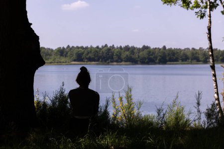 The photograph features the silhouette of a person seated in thoughtful solitude by the edge of a forest lake. The backdrop reveals a peaceful body of water encircled by trees, with sunlight