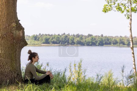 A young woman enjoys a moment of respite, leaning against a sturdy tree trunk, with a sprawling lake and lush greenery in her serene gaze. The peaceful environment and the distant treeline across the