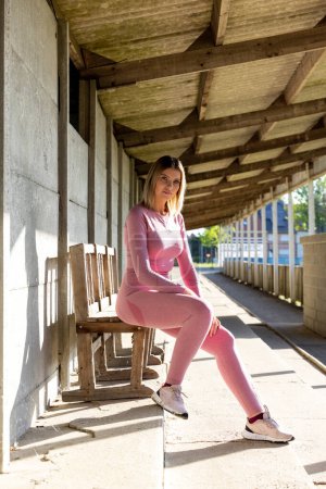 The image portrays a young woman seated on a rustic wooden bench in a spacious, shaded corridor, illustrating a moment of rest during her fitness routine. Dressed in a cohesive pink sportswear set
