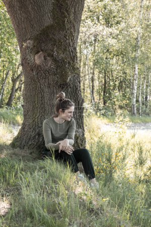 This image captures a young woman enjoying a quiet moment in the woods, seated at the base of a large tree with sunlight filtering through the leaves, creating a peaceful and reflective atmosphere