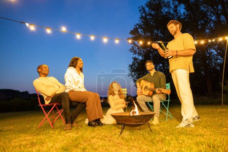 En el aire tranquilo de la noche, un grupo de amigos se relaja alrededor de un foso de fuego crepitante, con uno tocando la guitarra, todo bajo el suave destello de luces de cuerda en el fondo. Amigos disfrutando de un