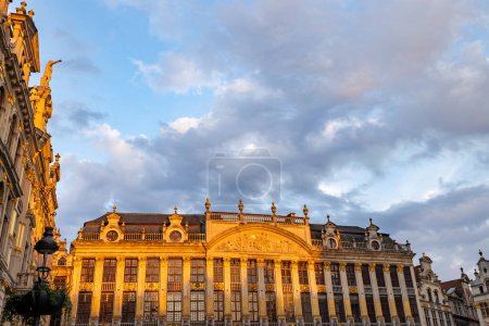 The historical guildhalls of Brussels Grand Place bask in the warm light of the golden hour, with ornate gilded facades illuminated against a backdrop of blue skies and wispy clouds. Golden Hour Glow