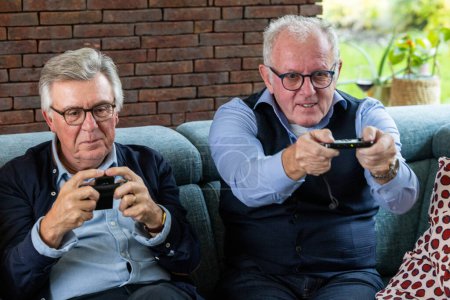 Zwei ältere Männer zeigen Konzentration und Spaß beim Spielen von Videospielen, bequem auf einer Couch sitzend. Eine Ziegelwand bietet eine warme Kulisse, die den modernen Zeitvertreib in einem heimeligen