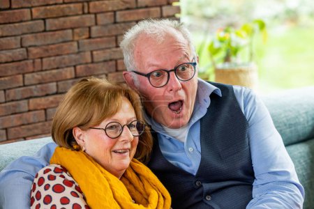 Un couple de personnes âgées partage un moment joyeux, avec l'homme exprimant sa surprise tandis que la femme sourit chaleureusement. Ils sont confortablement assis dans un cadre familial, avec un mur de briques en arrière-plan ajoutant
