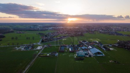 As the day ends, the setting sun casts a radiant backdrop over a small rural town. This aerial view showcases the juxtaposition of cultivated fields and residential areas, with the suns last light