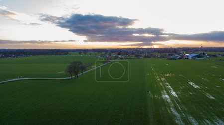 The evening sky graces a rural setting with sunbeams piercing through clouds, casting dramatic shadows over the green fields. This aerial shot captures the quiet beauty of the countryside as night