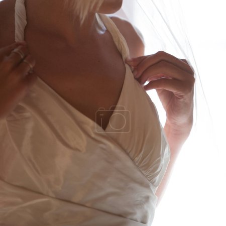 This intimate photograph focuses on the silhouette of a bride, capturing the grace and poise as she adjusts her gown. The soft backlighting creates a delicate interplay of light and shadow