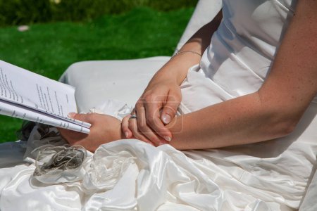This tender snapshot captures the hands of a bride, gently clasped over her lap, adorned with a wedding band. The simplicity of the white gown and the presence of wedding notes evoke the introspective