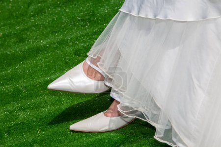 A brides classic pointed white wedding shoes peek from beneath the soft layers of a flowing gown against a lush green artificial turf, blending traditional bridal style with a touch of modernity