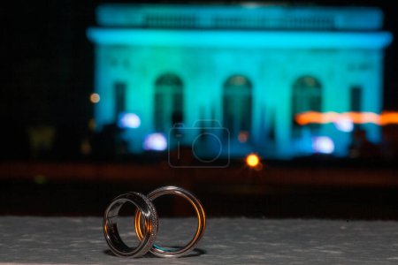 In the foreground, two wedding rings interlock with a bokeh backdrop of an elegant venue bathed in ambient night lights. This image captures the romance and solemnity of matrimonial symbols against a