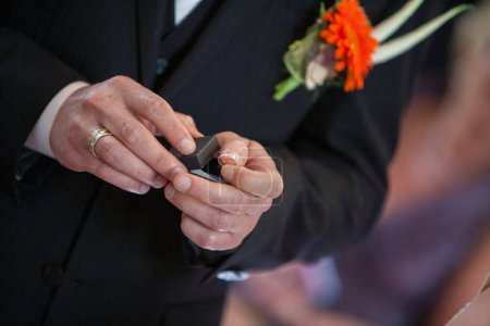 Un moment détaillé lors d'une cérémonie de mariage où un couple échange des anneaux, symbolisant leur engagement. Les mains des mariés tiennent soigneusement une boîte à bague alors qu'il se prépare à placer la bande sur son