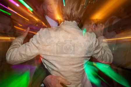 Capturing the energetic pulse of a dance floor, this image features the blurred movement of dancers with bursts of vibrant light streaks. The central figure in an elegant jacket becomes the focal