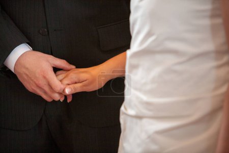 This close-up captures the tender moment where a bride places a wedding ring on the grooms finger. Their hands meet against the contrasting fabrics of the grooms suit and the brides dress, a