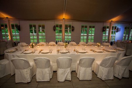 Une salle de banquet sophistiquée, prête pour les invités, avec des tables vêtues de linge blanc immaculé et ornées de délicats arrangements floraux. L'éclairage intime projette une lueur chaude, complétant le