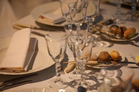 Eine Nahaufnahme eines raffinierten Speisesaals mit kristallklaren Gläsern, elegantem weißen Geschirr und fein gefalteten Servietten. Abgerundet wird das einladende Arrangement mit einer Auswahl an frisch gebackenen