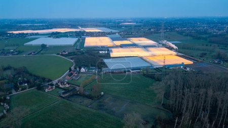 Téléchargez les photos : Duffel, Belgique, 20 mars 2024, Une perspective aérienne au crépuscule présente de vastes serres industrielles, leurs lumières brillantes comme des phares dans le paysage rural. Le contraste entre - en image libre de droit