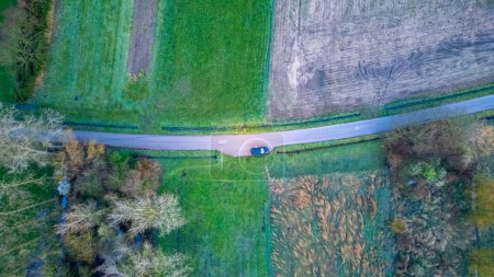 Esta fotografía aérea captura un vehículo solitario que viaja a lo largo de un sinuoso camino rural, rodeado por el mosaico de campos y árboles característicos del campo. La tranquilidad de la escena