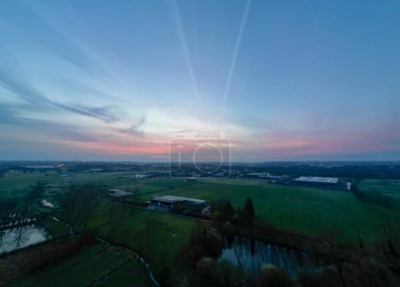 An aerial view captures the sprawling industrial greenhouses as day yields to night. The sunset casts a warm glow over the landscape, creating a striking balance between the industrial elements and