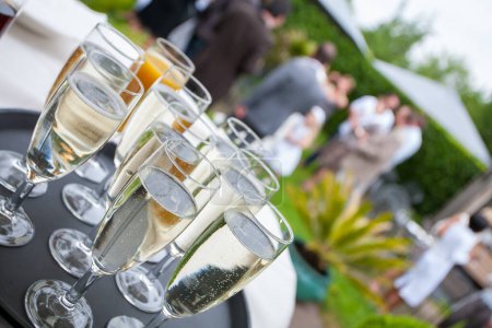 A tray of filled champagne flutes gleams in the foreground, inviting a celebratory toast at a garden party. The blurred background with guests hints at the social interactions and festive atmosphere