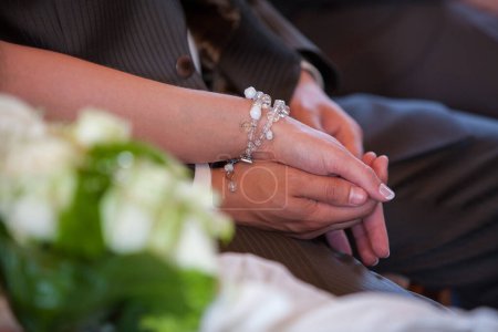 This photograph tenderly captures the intimate gesture of hand-holding at a formal event. The focus on the hands adorned with a delicate bracelet, alongside the soft blur of a floral arrangement