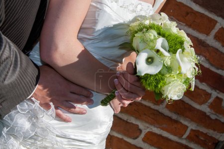 The image beautifully frames the clasped hands of a bride and groom against the backdrop of her elegant bridal bouquet and the timeless texture of a brick wall, capturing a moment of unity and