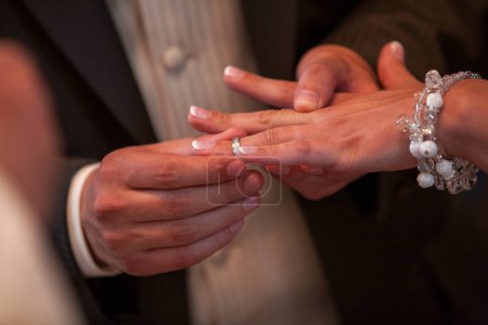Cette photographie capture un moment poignant au sein d'une cérémonie de mariage : le placement doux d'une alliance sur le doigt de la mariée. L'accent mis sur leurs mains sur le fond doux évoque le