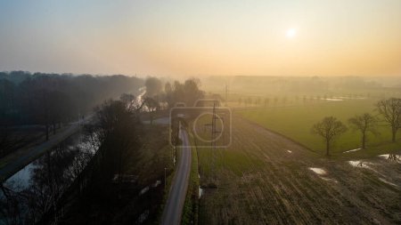 The morning sun casts a golden glow on a meandering road and canal, slicing through the fog-laden countryside. Sunrise Over Winding Road and Canal in a Misty Countryside Setting. High quality photo