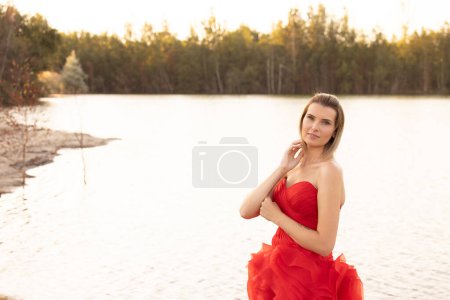 Inmitten der sanften Farbtöne eines ruhigen Seeufers in der Abenddämmerung strahlt eine Frau in einem kräftigen roten Kleid eine gelassene Eleganz aus, ihr nachdenklicher Ausdruck harmoniert mit dem ruhigen Wasser. Eleganz am See: Gelassene Frau