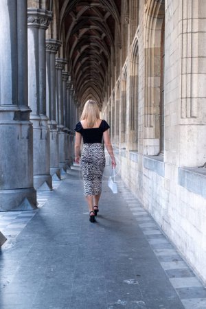 The photograph captures a young Caucasian woman with blonde hair, walking away from the viewer through a grand historical arcade. Her dark blue top and zebra-print skirt contrast with the stonework of