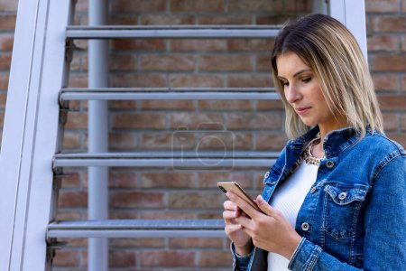 Eine moderne, fokussierte Frau interagiert mit ihrem Smartphone, lehnt an einem Metallgeländer vor Backsteinmauer-Hintergrund und veranschaulicht die Vernetzung des zeitgenössischen urbanen Lebens. Vernetzte Welt: Modern