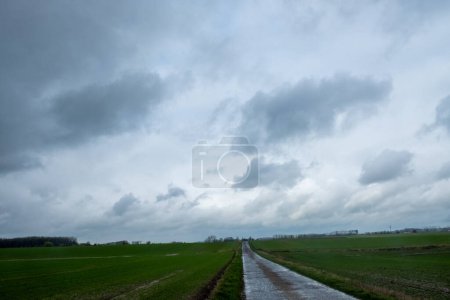 This dramatic landscape captures a rural road cutting through expansive green fields under brooding storm clouds. The contrast between the dark, heavy sky and the open field evokes the raw