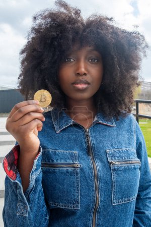 Eine junge schwarze Afroamerikanerin mit natürlichem, lockigem Haar hält eine Bitcoin-Münze in der Hand, ihr Gesichtsausdruck ist eine Mischung aus Neugier und Skepsis. In einer lässigen Jeansjacke steht sie draußen und symbolisiert