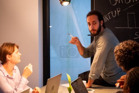 This image captures a moment from a strategic business meeting in a modern office setting. A man with a beard appears to be making a key point or presenting an idea to his colleagues, who listen