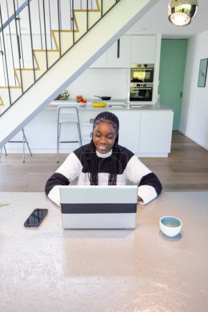 Ein helles und einladendes Bild einer glücklichen Frau mit geflochtenem Haar, die an einem geräumigen Küchentisch tief mit ihrem Laptop beschäftigt ist. Die moderne Küche Hintergrund schafft eine komfortable Umgebung, die verschmilzt