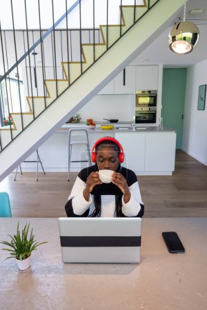 In einem Moment der Entspannung genießt eine Frau mit Kopfhörern eine warme Tasse Kaffee, während sie eine Pause von ihrer Arbeit am Laptop einlegt. Die geräumige und moderne Küche im Hintergrund, zusammen mit einer gut beleuchteten