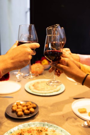 Eingefangen in einem warmen, umgebenden Licht, sieht man zwei Hände, wie sie Gläser Rotwein in einen Toast heben. Das Ambiente suggeriert ein gemütliches kulinarisches Erlebnis mit Häppchen auf dem Tisch, die den Betrachter in einen Moment einladen