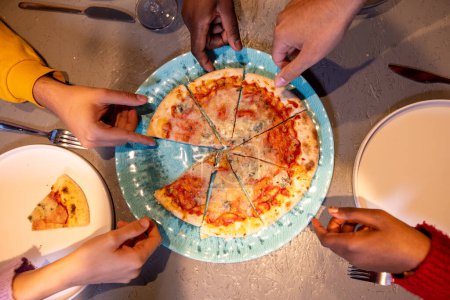 Ein Kopfschuss erfasst eine Gruppe von Freunden, als sie jeweils ein Stück einer frisch gebackenen Margherita-Pizza nehmen. Die Hände, die über dem Teller zusammenlaufen, symbolisieren Kameradschaft und die Freude am gemeinsamen Essen