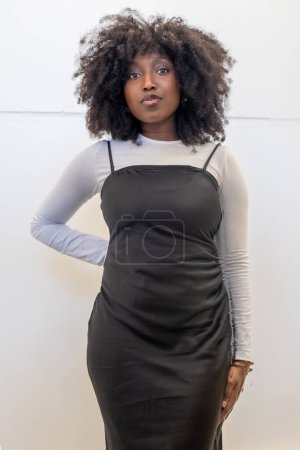 The image showcases a fashion savvy woman posing against a white background, her hair in a natural afro style. She is wearing a modern black slip dress over a fitted white long sleeve top, a trendy