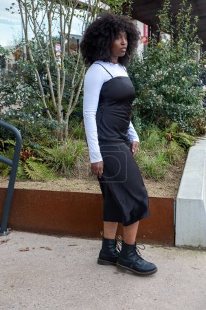 Cette image franche montre une femme élégante portant une longue robe noire sur un haut blanc à manches longues, associée à des bottes de combat noires. Ses cheveux bouclés et naturels encadrent son visage pendant qu'elle traverse une