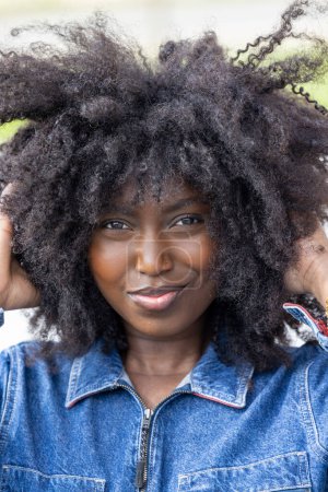 Das Porträt einer temperamentvollen Frau mit verspielter Afro-Frisur in Nahaufnahme, die der Kamera ein subtiles, selbstbewusstes Lächeln verleiht. Sie trägt eine Jeansjacke, die ihrem Look einen lässigen, aber kantigen Touch verleiht
