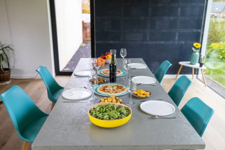 Das Bild zeigt einen modern gedeckten Esstisch mit gesunden Lebensmitteln, darunter frischem Salat und Pizza, die darauf warten, genossen zu werden. Die klare Linienführung des Tisches und die leuchtend blauen Stühle geben dem Raum eine